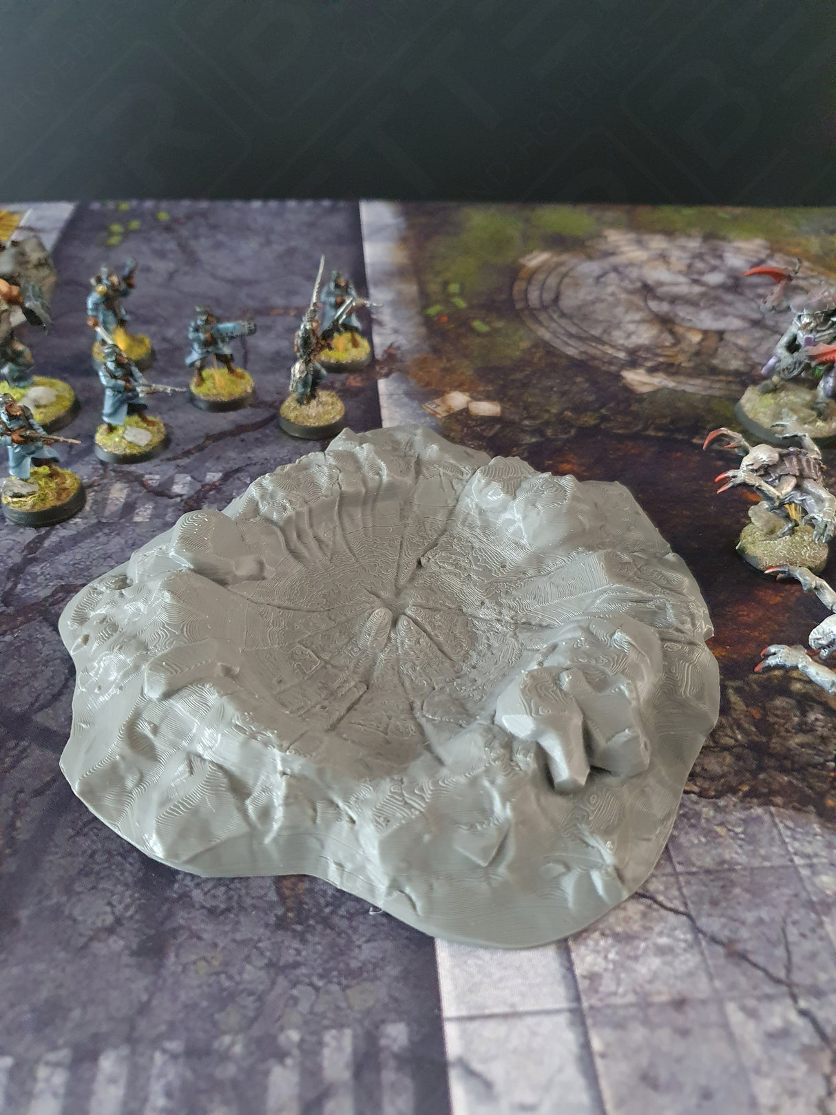Single grey 3D printed wargaming crater scatter terrain on a battle mat with miniatures for scale
